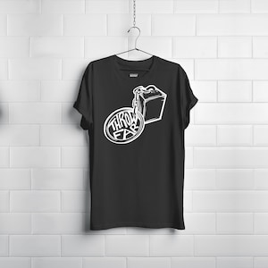 Puede incluir: Camiseta negra con un gráfico blanco. El gráfico presenta las palabras "THROW FAR" en un diseño circular, conectadas por una cadena a una caja cuadrada. La camiseta está colgada en una percha negra contra una pared de ladrillos blancos.