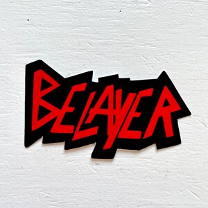 May include: A black sticker with the word "BEAVER" in red, stylized, jagged lettering. The sticker has a black border and is set against a white, textured background. The design is bold and eye-catching.