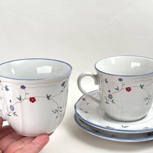 Op de afbeelding: Twee witte keramische theekopjes en twee bijpassende schoteltjes met een delicaat bloemmotief van kleine rode en blauwe bloemetjes op dunne groene ranken. Elk stuk heeft een gladde, geschulpte rand met een dunne blauwe lijn.