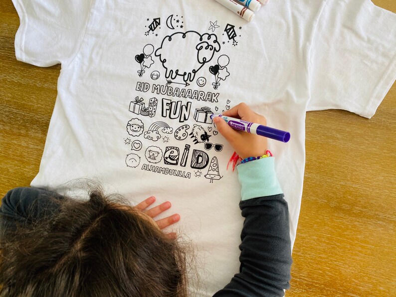 May include: A white t-shirt with black line drawings of a sheep, balloons, gifts, a rainbow, sunglasses, a trophy, a pizza slice, and other doodles. The text on the shirt reads 'Eid Mubaarak Fun Eid Alhamdulillah'.