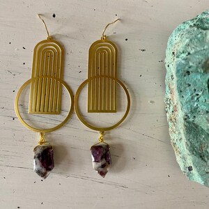 May include: Gold geometric earrings with a dangling red and white crystal. The earrings feature a circular design with a geometric pattern.