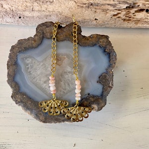 May include: A pair of gold earrings with a delicate chain and pink beads. The earrings feature a fan-shaped design at the bottom.