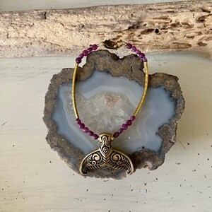 May include: A gold bracelet with a mermaid tail charm and purple beads. The bracelet is displayed on a piece of agate.