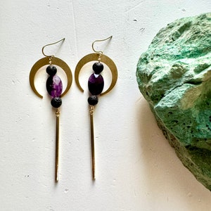 May include: A pair of gold crescent moon earrings with purple amethyst stones and black lava beads hanging from a gold bar.