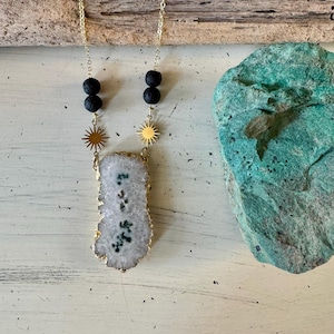 May include: A gold-colored chain necklace with a large, irregular white and green stone pendant. The necklace features black lava beads and gold sunburst charms. The pendant has a gold-colored edge. The necklace is displayed on a white surface.