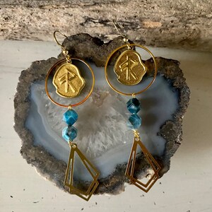 May include: Gold earrings with a blue gemstone and a gold charm with a rune symbol. The earrings are hanging from a gold hoop.