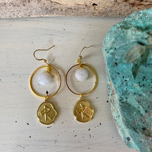 May include: Gold hoop earrings with a white stone and a gold charm with a rune symbol.