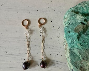 May include: A pair of gold earrings with a delicate chain and a black pearl dangling from each. The chain is adorned with small clear crystals.