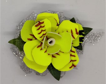 Puede incluir: Un arreglo floral con temática de softbol. Los pétalos de la flor son de color amarillo brillante con costuras rojas, que se asemejan a una pelota de softbol. Las hojas verdes y un respaldo plateado brillante completan el diseño. Un accesorio único con temática deportiva.