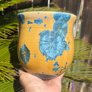 May include: A handmade ceramic vase with a yellow base and striking blue crystalline patterns. The vase is held in a hand, showcasing its unique shape and design. The exterior features a glossy finish, adding to its visual appeal.