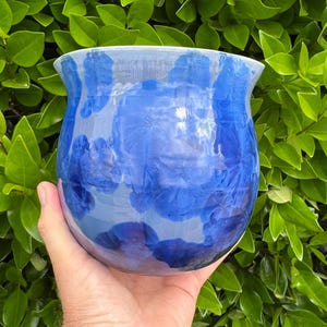 May include: A blue ceramic vase with a unique crystalline pattern. The vase has a rounded body and a slightly flared rim. The blue color is vibrant and the crystalline pattern adds visual interest. The vase is held in a hand against a backdrop of green foliage.