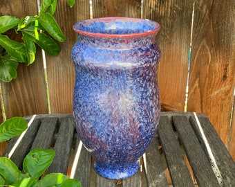 May include: A blue and purple ceramic vase with a speckled glaze. The vase has a rounded bottom and a wide opening at the top. It is sitting on a wooden table.