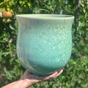 May include: A large, green ceramic planter with a speckled glaze. The planter has a rounded bottom and a wide opening.