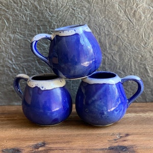 May include: Three blue ceramic mugs with white accents. The mugs are stacked on top of each other on a wooden surface.