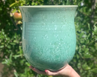 May include: A light green ceramic planter with a speckled glaze. The planter has a rounded bottom and a wide opening.