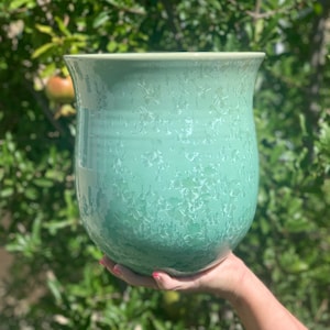 May include: A light green ceramic planter with a speckled glaze. The planter has a rounded bottom and a wide opening.