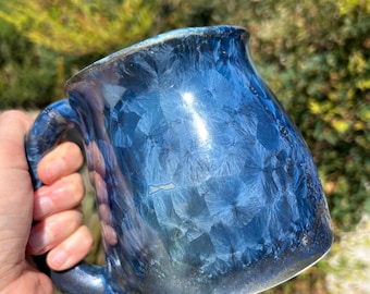 May include: A blue ceramic mug with a unique crystalline glaze. The mug has a rounded body and a handle.