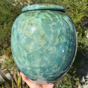May include: A round ceramic vessel with a glossy finish, featuring a crystalline glaze in shades of teal, turquoise, and pale green. The surface displays intricate, flower-like crystal patterns throughout, topped with a matching circular lid.