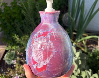 May include: A decorative ceramic soap dispenser with a white pump. The dispenser has a bulbous shape and is painted with a blend of red, purple, and blue hues. A white, crystalline pattern adorns the front, adding a unique touch to this bathroom accessory.