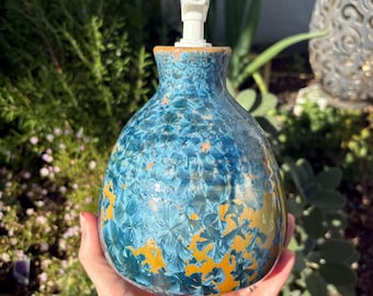 May include: A ceramic soap dispenser with a blue and gold crystalline glaze. The dispenser has a white pump and a rounded shape. The glaze has a textured appearance, with a pattern of blue crystals and gold accents. The dispenser is held in a hand.