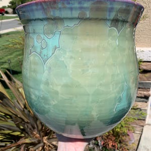 May include: A large, green ceramic planter with a mottled glaze. The planter has a wide, round base and a slightly flared rim. The glaze is a mix of light and dark green, with hints of blue and purple.
