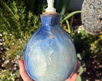 May include: A blue and white ceramic soap dispenser with a white pump. The dispenser has a bulbous shape with a textured, crystalline pattern. The top is a darker blue, transitioning to lighter shades. The dispenser is held in a hand.