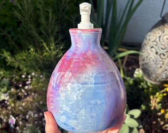 May include: A hand-held, ceramic soap dispenser with a white pump. The dispenser is painted with a blend of red, blue, and purple hues. The design has a textured, crystalline appearance, adding a unique aesthetic to the bathroom or kitchen.