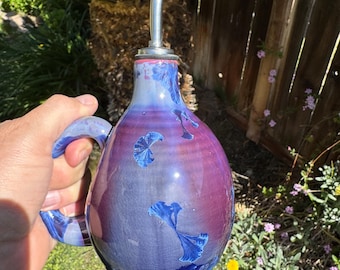 May include: A purple and blue ceramic oil and vinegar bottle with a silver spout and a handle. The bottle has a unique design with blue and white accents.