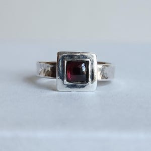 May include: A silver ring with a square, hammered setting. The setting holds a square, deep red gemstone. The band has a hammered texture. The ring is set against a white background.