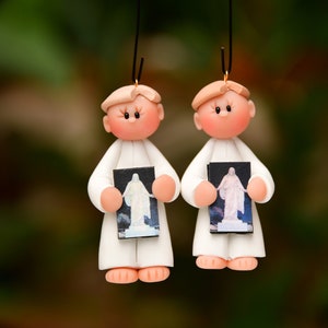 May include: Two white clay ornaments of children holding small framed pictures of a religious figure. The ornaments are hanging from a string.
