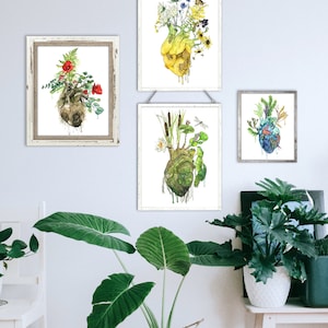 May include: Three framed watercolor paintings of anatomical hearts with different floral and plant arrangements growing from them. The hearts are yellow, green, and brown. The paintings are hanging on a white wall with a green plant in a woven basket in the foreground.