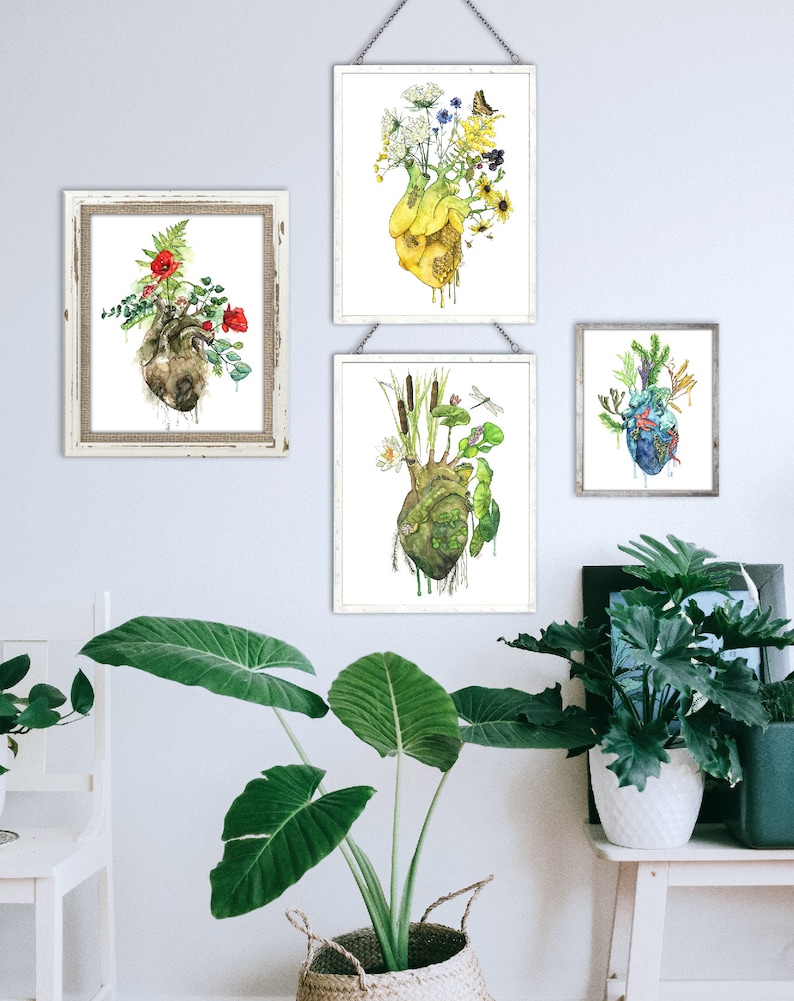 May include: Three framed watercolor paintings of anatomical hearts with different floral and plant arrangements growing from them. The hearts are yellow, green, and brown. The paintings are hanging on a white wall with a green plant in a woven basket in the foreground.