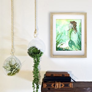 May include: A watercolor painting of a mermaid with long brown hair, wearing a white top and a green tail. The painting is framed in a light wood frame and hanging on a white wall. There are two hanging glass globes with green plants in them.