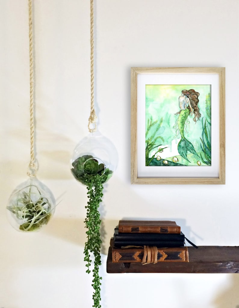 May include: A watercolor painting of a mermaid with long brown hair, wearing a white top and a green tail. The painting is framed in a light wood frame and hanging on a white wall. There are two hanging glass globes with green plants in them.