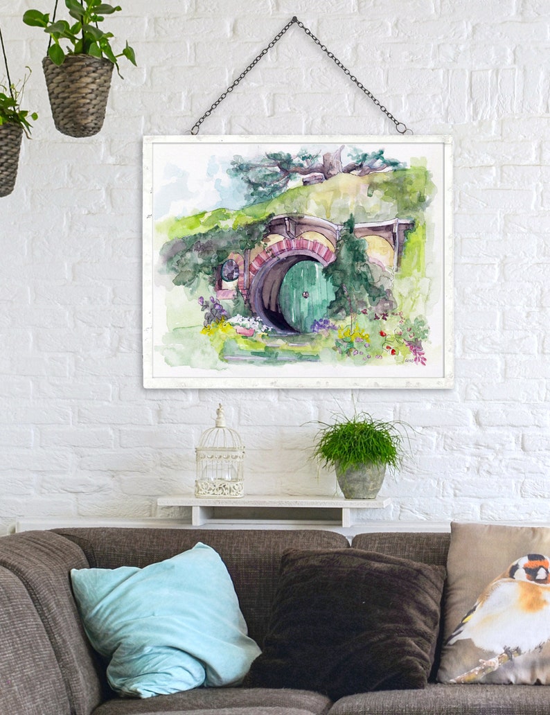 May include: A watercolor painting of a hobbit hole with a green door. The hole is surrounded by lush greenery and flowers. The painting is framed in white and hanging on a white brick wall.