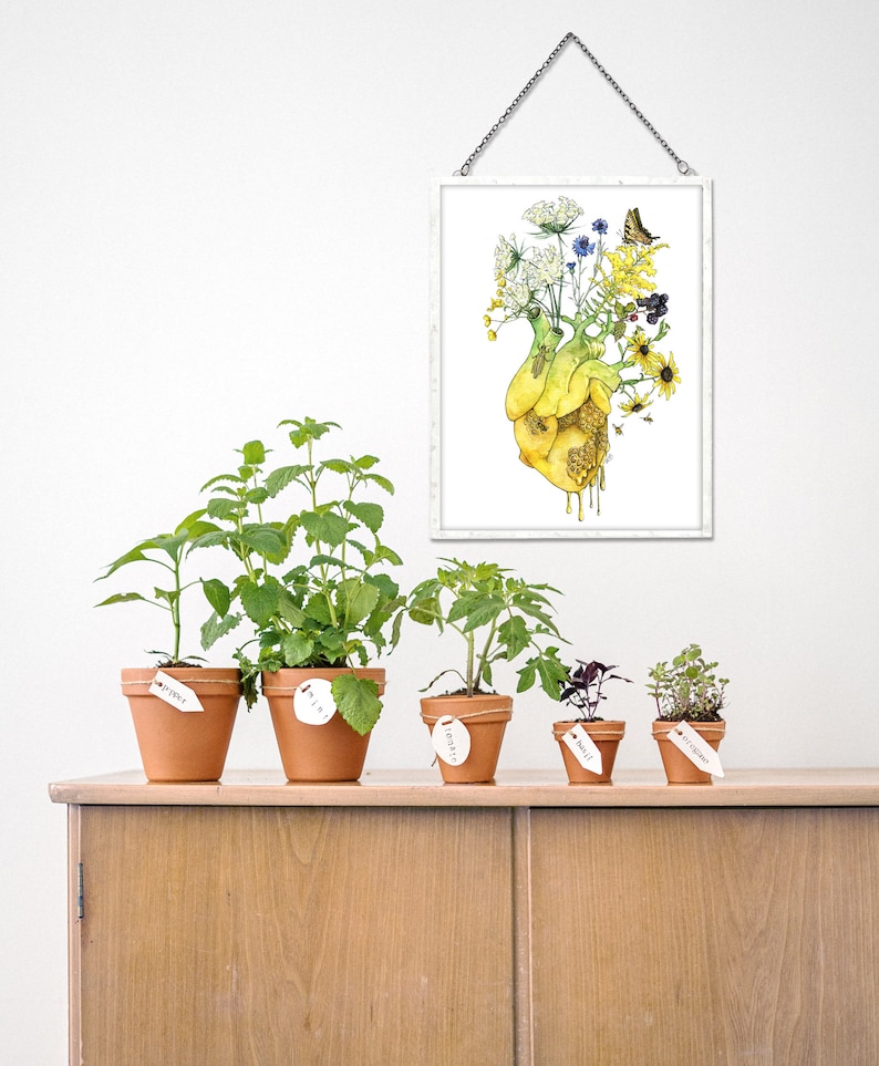 May include: Five potted plants with terracotta pots and white labels. The plants are arranged on a wooden shelf. The labels are handwritten with the names of the plants: "Thyme", "Mint", "Tomato", "Basil", and "Rosemary". A framed print of a yellow heart with flowers and a butterfly hangs on the wall above the shelf.