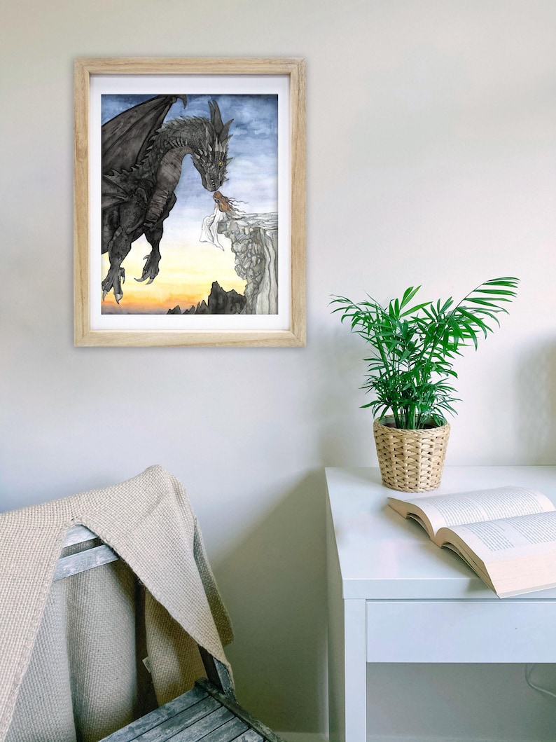 May include: A framed watercolor painting of a black dragon with a woman standing on a cliff. The dragon is flying towards the woman and appears to be breathing fire. The background is a sunset with mountains in the distance.