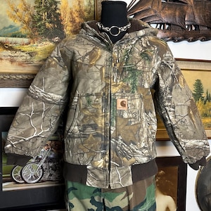 Puede incluir: Una chaqueta Carhartt con estampado de camuflaje con capucha, bolsillos delanteros y dobladillo y pu&ntilde;os acanalados marrones. La chaqueta tiene cierre de cremallera y un peque&ntilde;o parche con el logotipo de Carhartt en el bolsillo delantero. La chaqueta se muestra en un maniqu&iacute;.