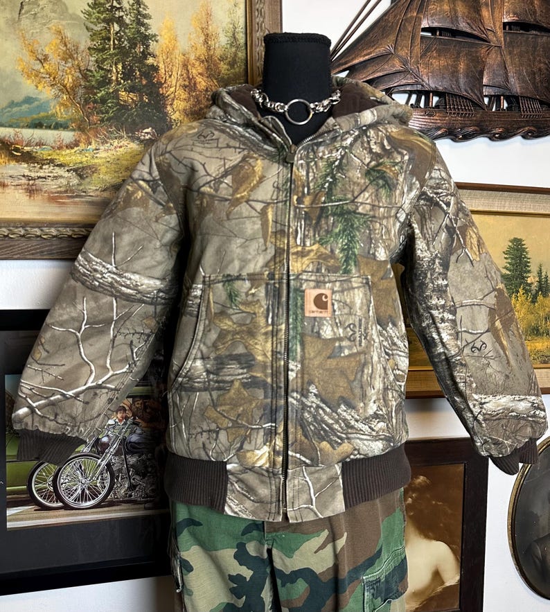 Puede incluir: Una chaqueta Carhartt con estampado de camuflaje con capucha, bolsillos delanteros y dobladillo y pu&ntilde;os acanalados marrones. La chaqueta tiene cierre de cremallera y un peque&ntilde;o parche con el logotipo de Carhartt en el bolsillo delantero. La chaqueta se muestra en un maniqu&iacute;.
