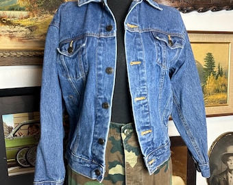 May include: A mid-wash blue denim jacket with button fastenings and pockets. The jacket is displayed on a black mannequin, paired with camouflage cargo trousers. The jacket has a classic collar and long sleeves.