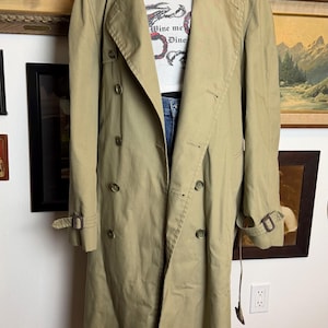 May include: Khaki trench coat with a double-breasted design, button closures, and a belt. The coat is paired with a white graphic tee that says "Wine me Dine". The coat has a classic, timeless style.