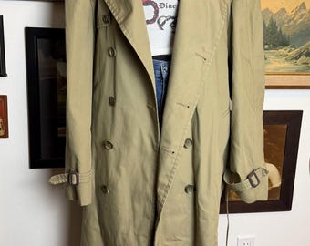 May include: A khaki trench coat with a double-breasted design, button fastenings, and a belt. The coat is worn with a white graphic tee that says "Wine me Dine". The coat has a classic, timeless style.