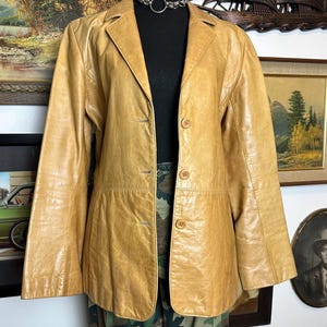 May include: A tan leather blazer with a classic notched collar and button closure. The jacket has a tailored fit and long sleeves. The blazer is displayed on a black mannequin, paired with camouflage pants, and a silver chain necklace.