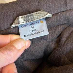 Puede incluir: Primer plano de una etiqueta de prenda marr&oacute;n con el nombre de la marca "Carhartt" en letras blancas. La etiqueta tambi&eacute;n indica la talla M (10-12) y "Made in Vietnam". La prenda parece acolchada.