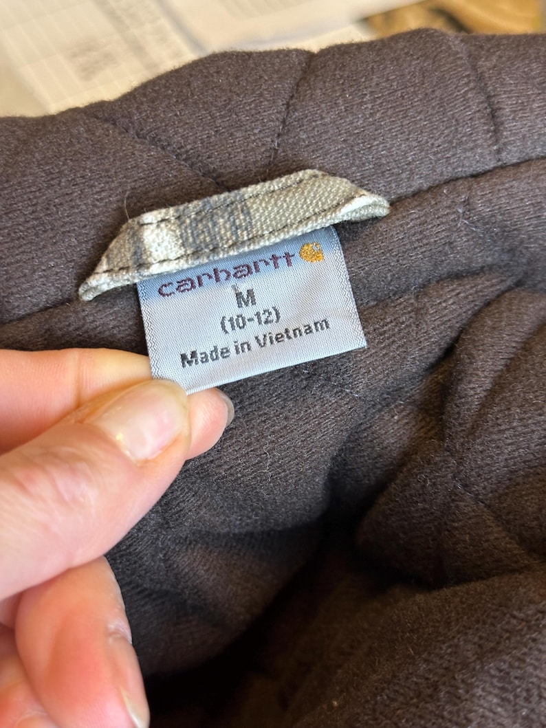 Puede incluir: Primer plano de una etiqueta de prenda marr&oacute;n con el nombre de la marca "Carhartt" en letras blancas. La etiqueta tambi&eacute;n indica la talla M (10-12) y "Made in Vietnam". La prenda parece acolchada.