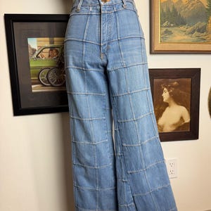 May include: Light blue denim jeans with a unique patchwork design. The jeans have a high waist and a wide leg, with a grid-like pattern created by the stitching. The top features a white tank top with the text "Dine me".