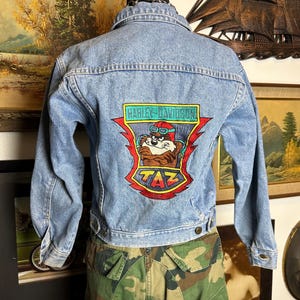 May include: Light blue denim jacket with a Harley-Davidson Taz patch on the back. The patch features Taz the Tasmanian Devil wearing goggles and a helmet. The jacket has a button closure and long sleeves. The model is wearing camouflage pants.