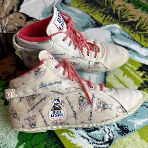 May include: Pair of vintage Bud Light high-top sneakers. The canvas shoes feature a repeating pattern of the Bud Light logo and the Spuds MacKenzie mascot. They have red laces and interior lining.