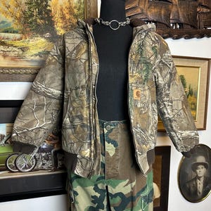 Puede incluir: Conjunto de chaqueta y pantal&oacute;n con estampado de camuflaje sobre un maniqu&iacute;. La chaqueta tiene capucha y dobladillo acanalado marr&oacute;n. Los pantalones son de estilo cargo. Se lleva un collar de gargantilla plateado alrededor del cuello del maniqu&iacute;.