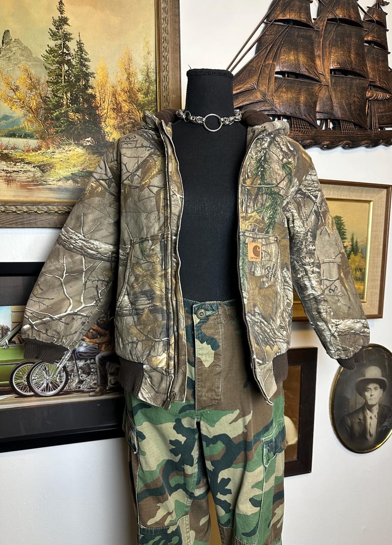 Puede incluir: Conjunto de chaqueta y pantal&oacute;n con estampado de camuflaje sobre un maniqu&iacute;. La chaqueta tiene capucha y dobladillo acanalado marr&oacute;n. Los pantalones son de estilo cargo. Se lleva un collar de gargantilla plateado alrededor del cuello del maniqu&iacute;.